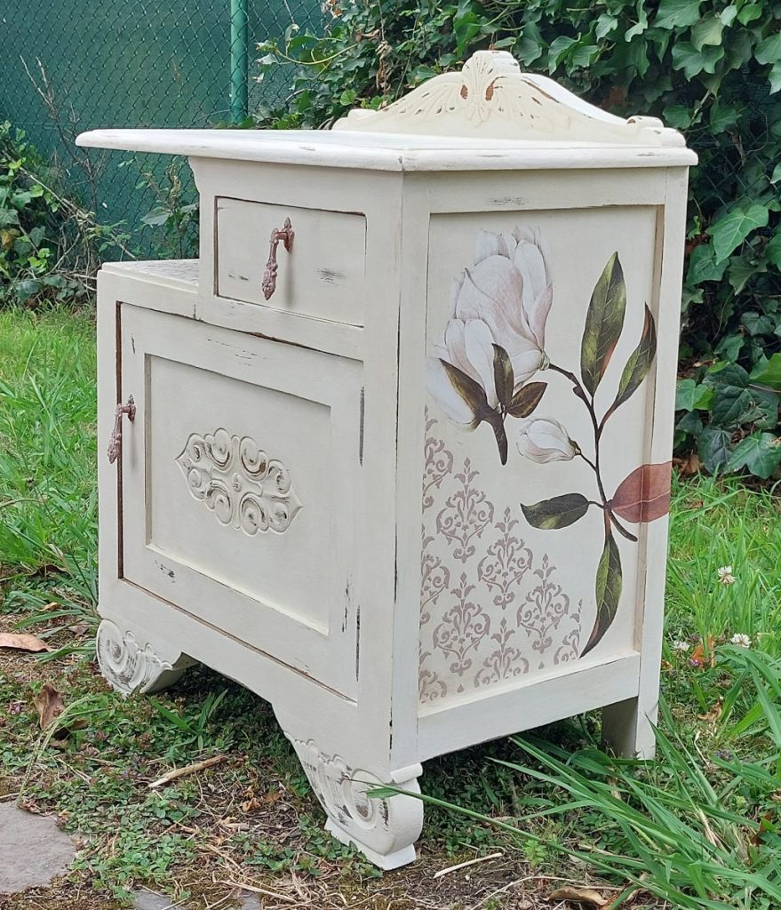Mesita de noche antigua pintada de blanco con transferencias de magnolias en los laterales, desgastes y estarcidos decorativos.