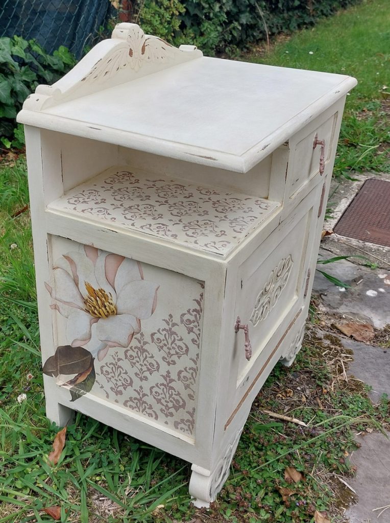 Mesita de noche antigua pintada de blanco con transferencias de magnolias en los laterales, desgastes y estarcidos decorativos.