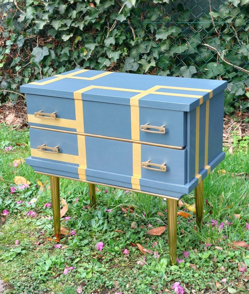 Mesilla de dos cajones con tiradores originales de metal dorado. Está pintada de azul con líneas doradas atravesándolo. Tiene patas doradas también.