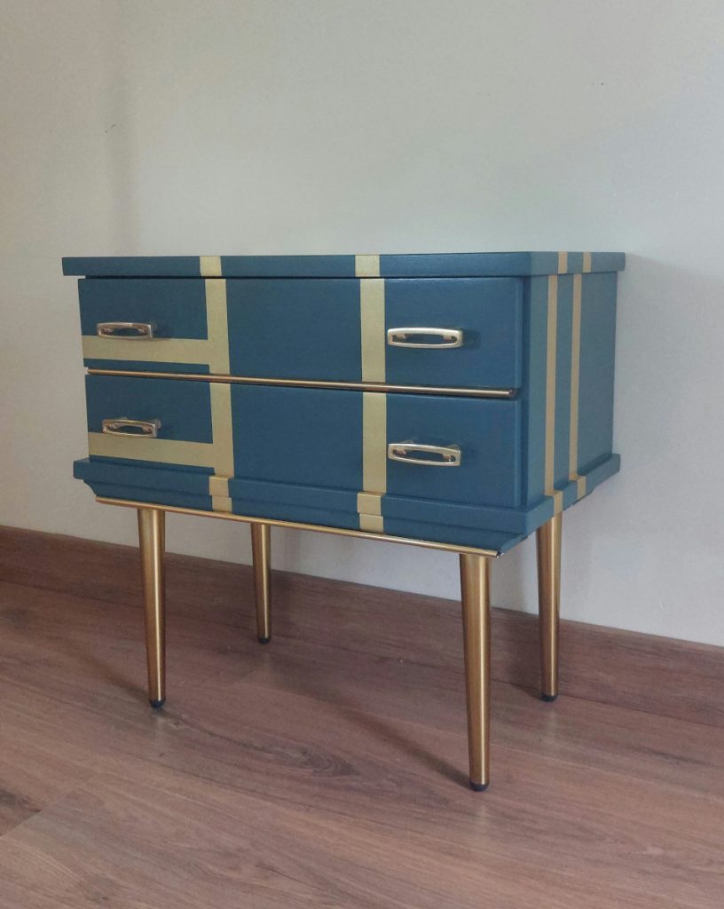 Mesilla de dos cajones con tiradores originales de metal dorado. Está pintada de azul con líneas doradas atravesándolo. Tiene patas doradas también.