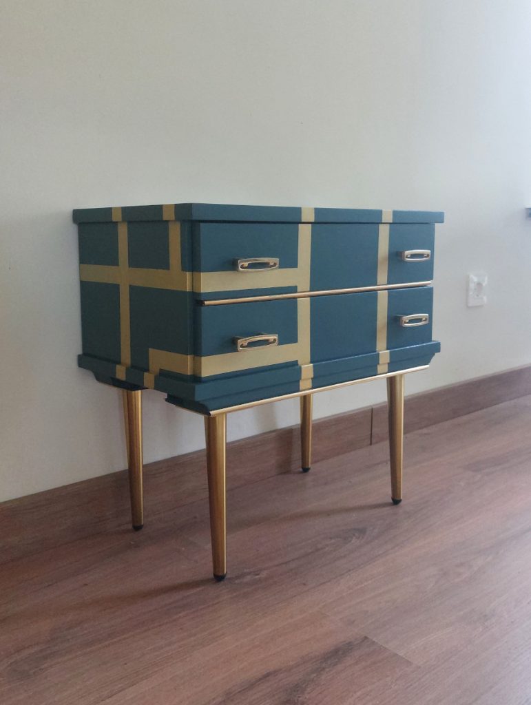 Mesilla de dos cajones con tiradores originales de metal dorado. Está pintada de azul con líneas doradas atravesándolo. Tiene patas doradas también.